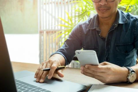 Man's hands using cell phone while working with laptop on wooden desk. Stock Photos