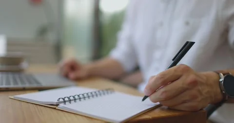 Man's hands writing notes in notebook with black ball pen sitting with laptop at Stock Footage 160771188