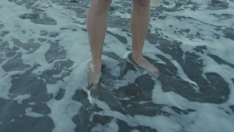 A man's legs are washed by the waves of the sea, who stands on the black sand Stock Footage 307755869