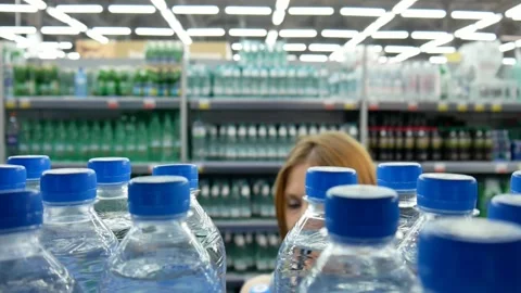 Many bottles of potable water on a store shelf and a young woman chooses one Vidéo 247213221