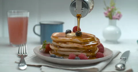 Maple syrup is poured over a stack of pancakes with fruit and butter on table in Stock Footage 199460299