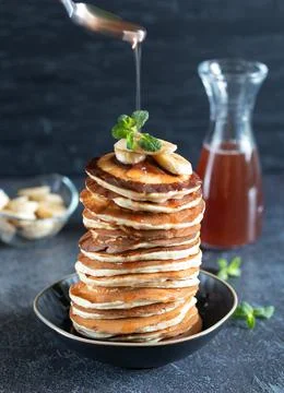 Maple syrup pours from a spoon onto a stack of pancakes with bananas and mint Stock Photos