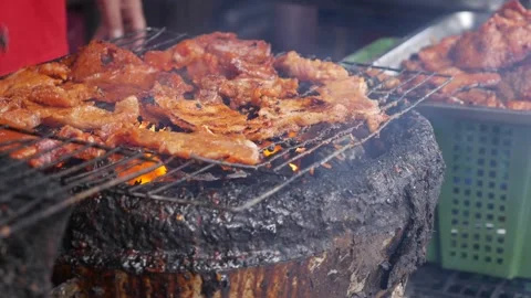 A market vendor grills pieces of meat over hot charcoal near a food stall with c Stock Footage 338001353