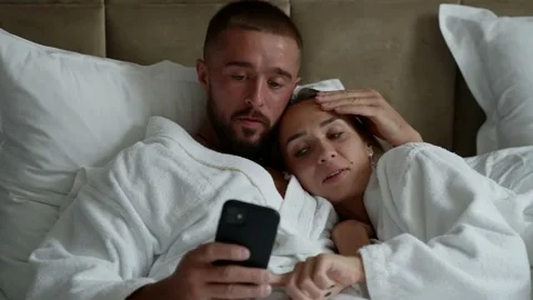 Married couple is lying in bed and watching pictures in smartphone, man and Видео 163756475