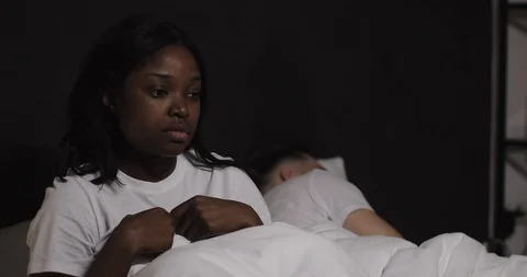 Married multi-ethnic couple lying in bed at night. Worried woman sitting on bed Stock Footage 111136165