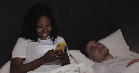 Married multi-ethnic couple lying in bed at night. Woman using smartphone Stock Photos