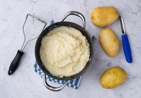 Mashed potatoes in a pan with potatoes and utensils over white marble surface Stock Photos
