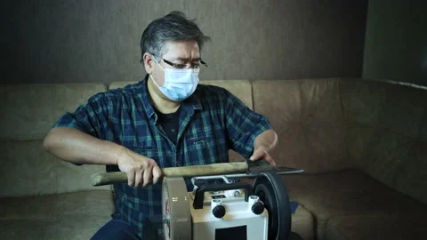 Masked asian man sharpens axe on a grinder Stock Footage 143673732