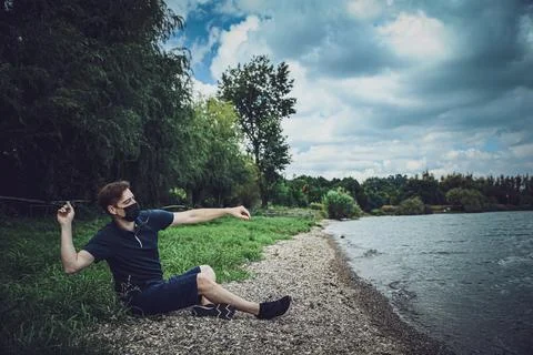 Masked guy is sitting and throwing a stone to lake Stockfoto's