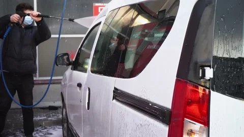 Masked man in black jacket washes his white car, douses it with water from hose Stock Footage 133175920