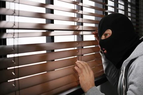 Masked man spying through window blinds indoors. Criminal offence Stock Photos