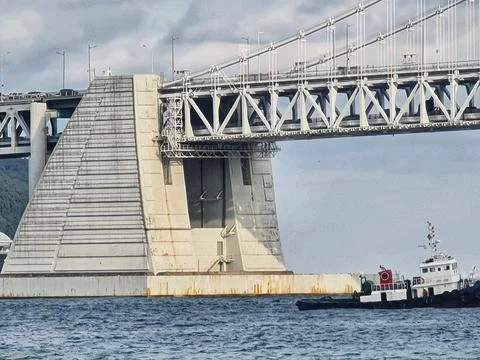A massive bridge support extends over calm water with a small tugboat nearby  Stock Photos