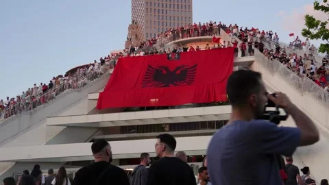 Massive crowd shouting enthusiastically below large Albanian flag hanging on Stock Footage 338033064