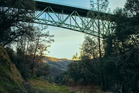 The massive metal bridge structure rises above green trees and mountains, cre Stock Photos