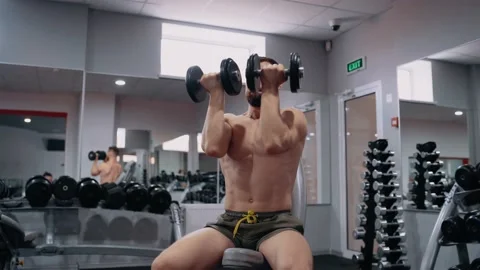 The massive, sporty man performs lifting the dumbbells, in the gym, in a sitting Video stock 156477808