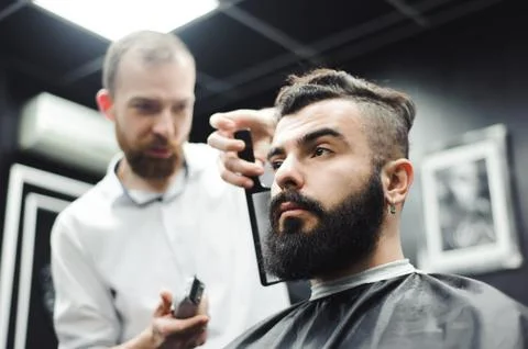 Master cuts hair and beard of men in the barbershop, hairdresser makes hairstyle Stock Photos