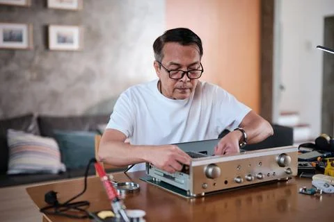 Mature Asian man disassembling audio equipment for repair, home DIY workshop. Stock Photos