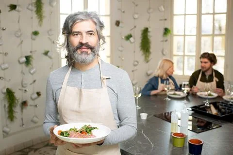 Mature bearded man in pullover and apron holding plate with selfmade vegetable Stock Photos