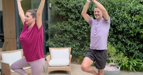 Mature couple doing tree-pose on patio over green yoga mats woman lifting man Stock Footage 341852379
