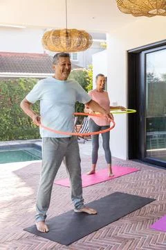 Mature couple exercising using orange and yellow hula hoops on covered patio Stock Photos