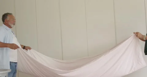 Mature couple shaking, pulling pink sheet across bed after laundry with basket, Stock Footage 337540813
