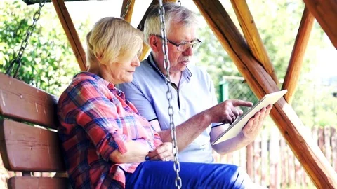 Mature couple talking and using tablet computer sitting on the swing in the gard Video stock 78764255
