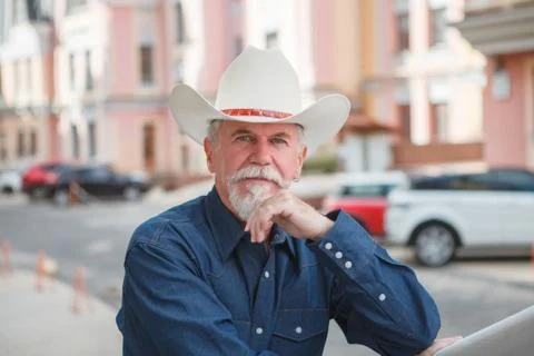 A mature cowboy in a hat, jeans and a denim shirt looks at the camera. On ope Stock Photos