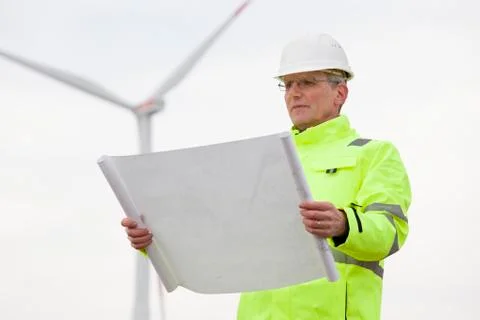 Mature engineer standing in front of a wind turbine with blueprint in his han 写真素材