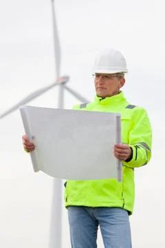 Mature engineer standing in front of a wind turbine with blueprint in his han 스톡 사진