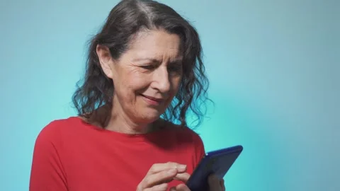 Mature female smiling while texting on smartphone over blue background Stock Footage 240132423