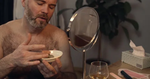 Mature good looking man applying facial mask while sitting in front of mirror Stock Footage 136476980