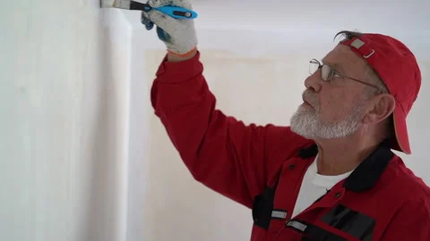 Mature gray-bearded man in a red cap and glasses paints the wall with a white Stock Footage 165456960