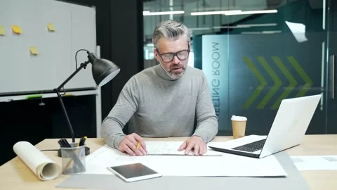 A mature gray haired bearded man in glasses is drawing a project on paper while  Stock Footage 232576643