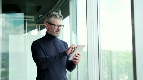 Mature gray haired bearded man wearing glasses uses tablet while standing near  Stock Footage 232578449