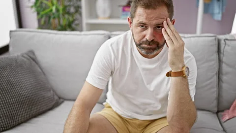 Mature hispanic man sitting thoughtfully on a grey sofa indoors, portraying.. Видео 272983229