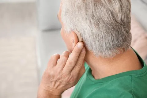 Mature man adjusting hearing aid at home, closeup Stock Photos