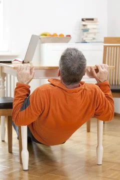 Mature man doing pull-ups at home under a dining table with laptop Stock Photos