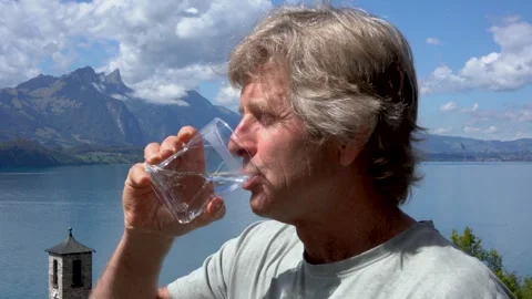 Mature man drinking fresh water from glass with mountain lake view in background Video stock 343727426