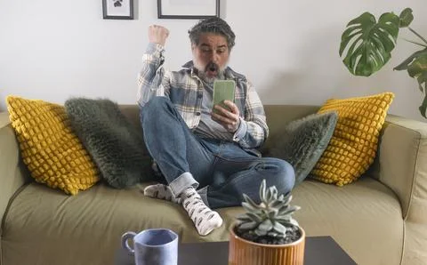 Mature man expressing excitement while using smartphone, raising fist in ce.. Stock Photos