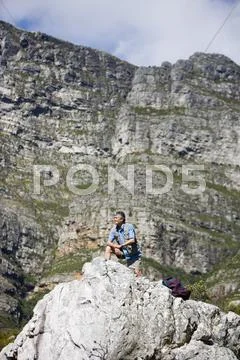 Mature man hiking on mountain trail, crouching on rock, looking at scenery, s Stock Photos