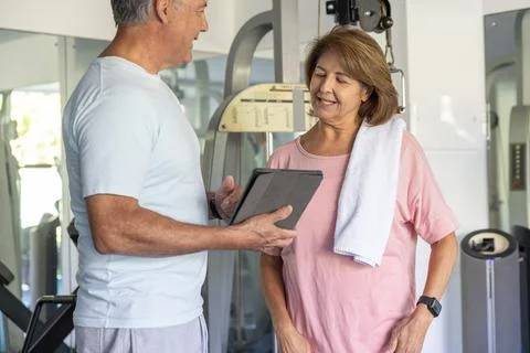 Mature man holding tablet instructing senior woman wearing pink tee with towel Stock Photos