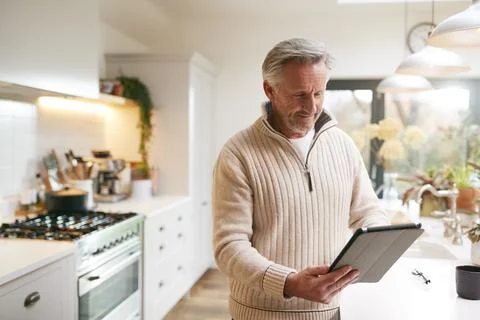 Mature Man In Kitchen Working From Home Using Digital Tablet Fotos de archivo