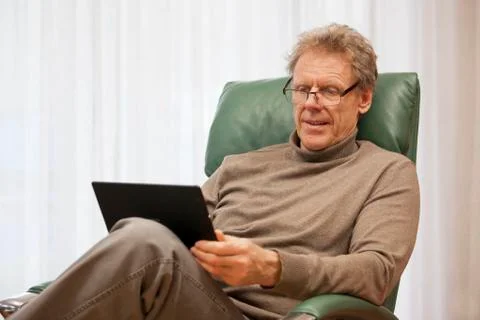 Mature man looking at a tablet while sitting in a chair in a bright living ro Stock Photos