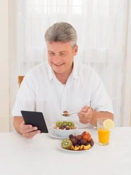 Mature man reading book while eating breakfast Stock Photos