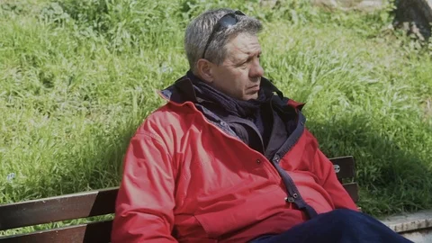 Mature Man in a red Jacket on a bench in the Park takes the phone out of pocket Vidéo 100947497