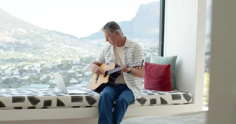 Mature man settling on window bench, tuning acoustic guitar, glancing at laptop Stock Footage 336484139