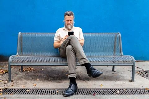 Mature man sitting on a bench using mobile phone. Blue background. Copy space. Stock Photos