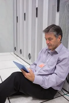 Mature man sitting on raised floor among server racks studying tablet with ID Stock Photos
