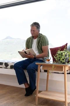 Mature man sitting on window bench at home, wearing green shirt, writing with Stock Photos