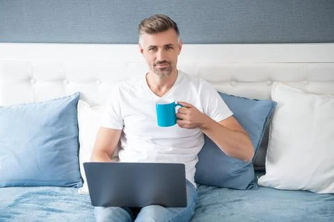 Mature man smile using computer in bed with coffee Stock Photos